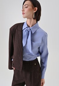 Camicia azzurra con colletto a farfalla, abbinata a un blazer marrone scuro con sottili righe verticali. Tessuto liscio, vestibilità strutturata e pantaloni con vita elastica.