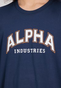 Marineblauwe katoenen T-shirt met een ronde hals, voorzien van een groot "ALPHA" logo in het wit, omrand met oranje, en "INDUSTRIES" in kleinere witte tekst.