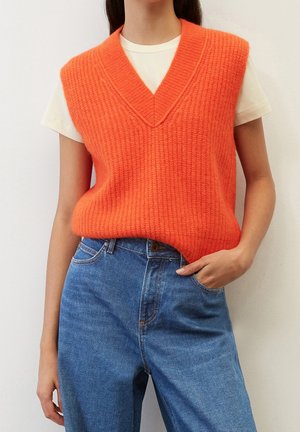 Frau trägt eine leuchtend orange ärmellose Strickweste über einem weißen T-Shirt, kombiniert mit hoch taillierten blauen Jeans, eine Hand in der Tasche.