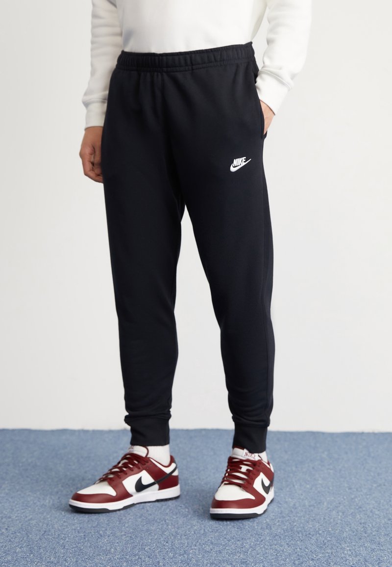 Person klädd i svarta Nike sweatpants och röda, vita och svarta Nike-skor som står på en blå matta med en hand i fickan.