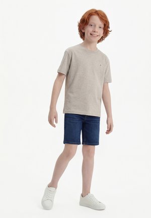 Băiat cu păr roșcat zâmbind, purtând un tricou gri deschis, pantaloni scurți de denim albastru închis și adidași albi, stând pe un fundal alb.