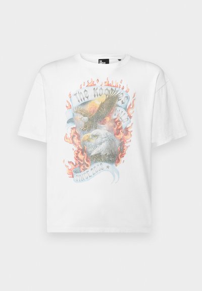 Camiseta de algodón blanca con un gráfico de dos águilas rodeadas de llamas y una cinta que dice "Desde 2000", en colores vibrantes.