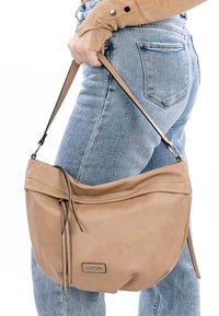 Beige leren handtas met een ontspannen ontwerp, verstelbare schouderriem en zichtbaar logo. Bevat een gevouwen bovenkant en twee hangaccessoires.