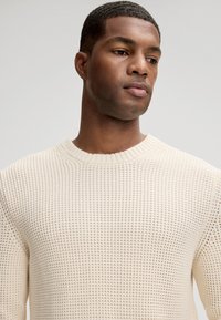 Sweter z dzianiny w kolorze ecru, o fakturowanym splocie, z okrągłym dekoltem i ściągaczami na mankietach. Przylegający przy szyi, z luźną sylwetką.