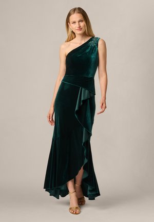 Femme en robe asymétrique en velours vert forêt à une épaule, avec un ourlet à volants et des talons dorés à brides, sur fond uni.