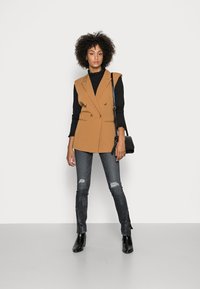 Een tan dubbelgeknoopte vest met een zwarte coltrui, gecombineerd met versleten donkere jeans en enkelboots, staat tegen een witte achtergrond.