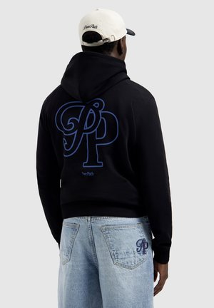 Mann trägt weiße Kappe, schwarzen Hoodie mit großem blauem "PP"-Logo und "Pure Path"-Schriftzug auf dem Rücken sowie hellblaue Jeans mit passender "PP"-Stickerei.