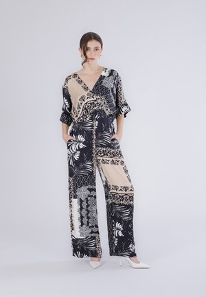 Nainen seisoo mustavalkoisessa, kuviollisessa leveälahkeisessa haalariasussa, jossa on V-pääntie ja kädet taskuissa. Hänellä on valkoiset korkokengät, ja tausta on yksivärinen.