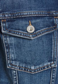 Blue denim fabric with a chest pocket featuring a silver button and orange stitching. Textured surface with slight fading and wear details.