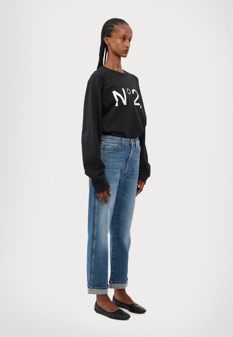 Svart hettegenser med hvit "N°2" grafikk, sammen med lys blå rettbente jeans og svarte flate sko. Bomullsmaterialet er synlig i genseren.