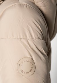 Chaqueta acolchada beige con un parche del logo redondeado de Calvin Klein en la manga, textura suave con patrón acolchado y costuras cosidas.