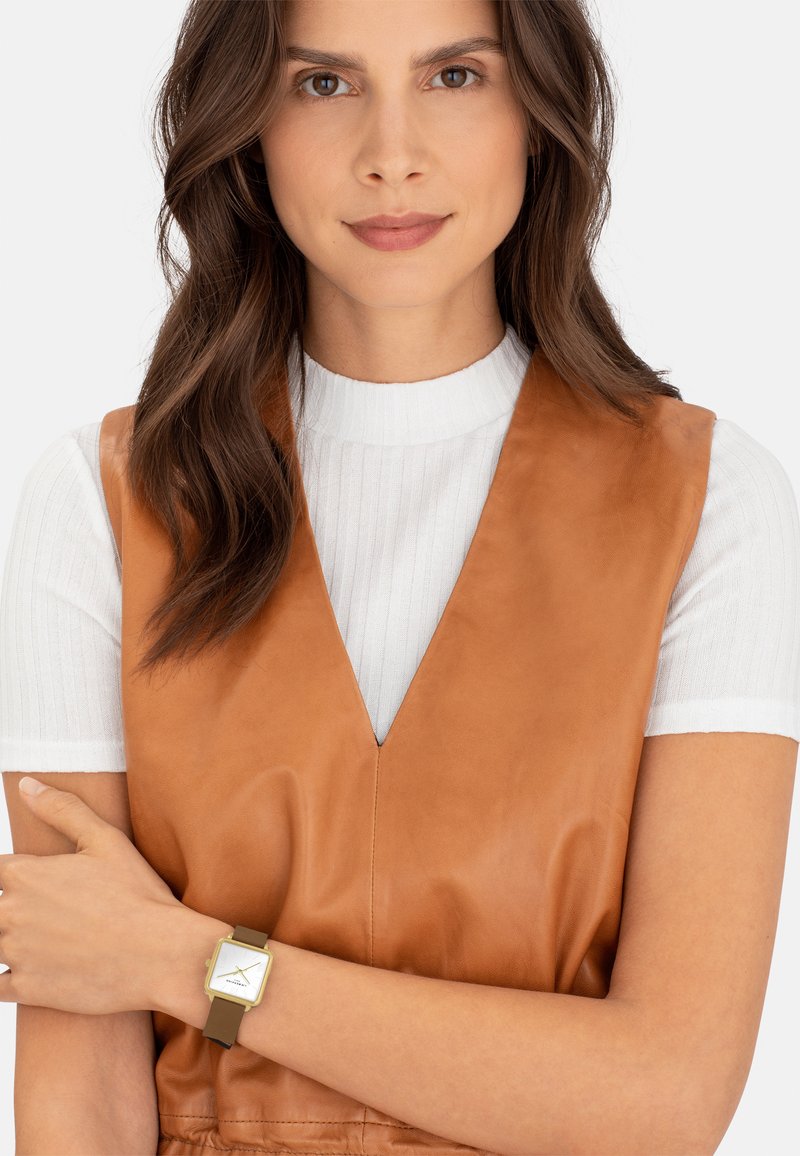 Bruine leren vest met een diepe V-hals over een witte geribbelde shirt; draagt een vierkante horloge met een groene band en gouden accenten.