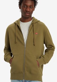 Olivenfarget zip-up hettegenser med snøring, frontlommer og en liten rød logo på venstre bryst. Mykt stoff med avslappet passform.