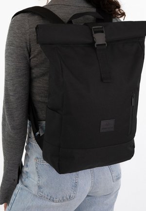 Person trägt einen großen schwarzen Rucksack mit Rollverschluss und Schnallen, seitlicher Reißverschlusstasche, grauem Pullover und hellblauen Jeans.