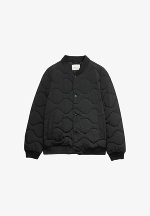 Man draagt een zwarte doorgestikte bomberjas, zwart shirt, witte broek, zwarte riem en zwarte leren schoenen, staand tegen een effen achtergrond.