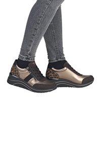 Ben i grå skinny jeans iført bronze- og sorte sneakers med leopardmønstrede hæle og sidelommer, stående krydset på hvid baggrund.