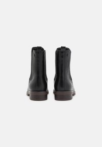 Un par de botas de piel negras hasta el tobillo con tiradores y tacones bajos de madera marrón, vistas desde atrás sobre un fondo blanco.