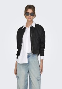 Black bomber jacket with ribbed cuffs and zipper, worn over a white collared shirt, paired with light blue baggy jeans and black sunglasses.