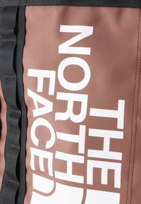 Κοντινό πλάνο καφέ υφάσματος με λευκή επιγραφή "THE NORTH FACE" και διπλανό μαύρο ιμάντα με ραμμένα θηλάκια.