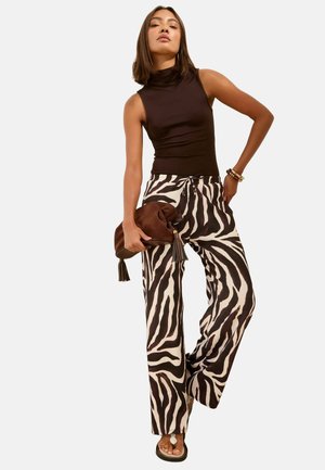 Woman wearing sleeveless black turtleneck top, zebra-print wide-leg pants, holding brown suede tassel clutch, and wearing sandals.
