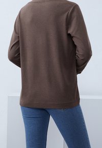 Camiseta de manga larga marrón hecha de material suave, con un escote redondeado y un corte relajado. Se lleva con jeans azules, mostrando un estilo casual.