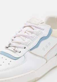 Baskets en cuir blanc avec des accents bleu clair, des lacets blancs, un bout perforé et une étiquette métallique de marque sur les lacets.