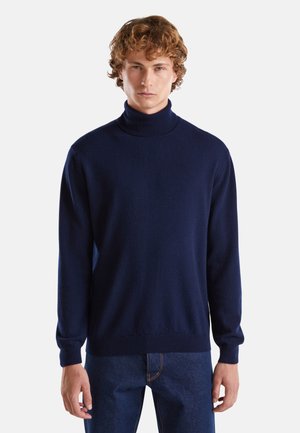 Maglione blu navy a collo alto, realizzato in materiale morbido, con maniche lunghe e polsini e orlo a costine. Indossato con jeans blu scuro.