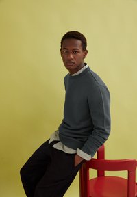 Sweatshirt gris avec un col rond sur une chemise à col gris clair, associé à un pantalon sombre, assis sur une chaise rouge devant un fond jaune.