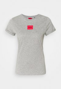 Camiseta de algodón gris con mangas cortas, presenta un parche rectangular rojo con el logotipo "HUGO" en el centro, escote redondo, textura suave.
