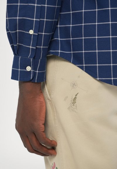 Temno modra karo srajca z belimi mrežastimi črtami, gumbi na manšetah, v kombinaciji s bež hlačami z vezenimi detajli in motivom kompasa.
