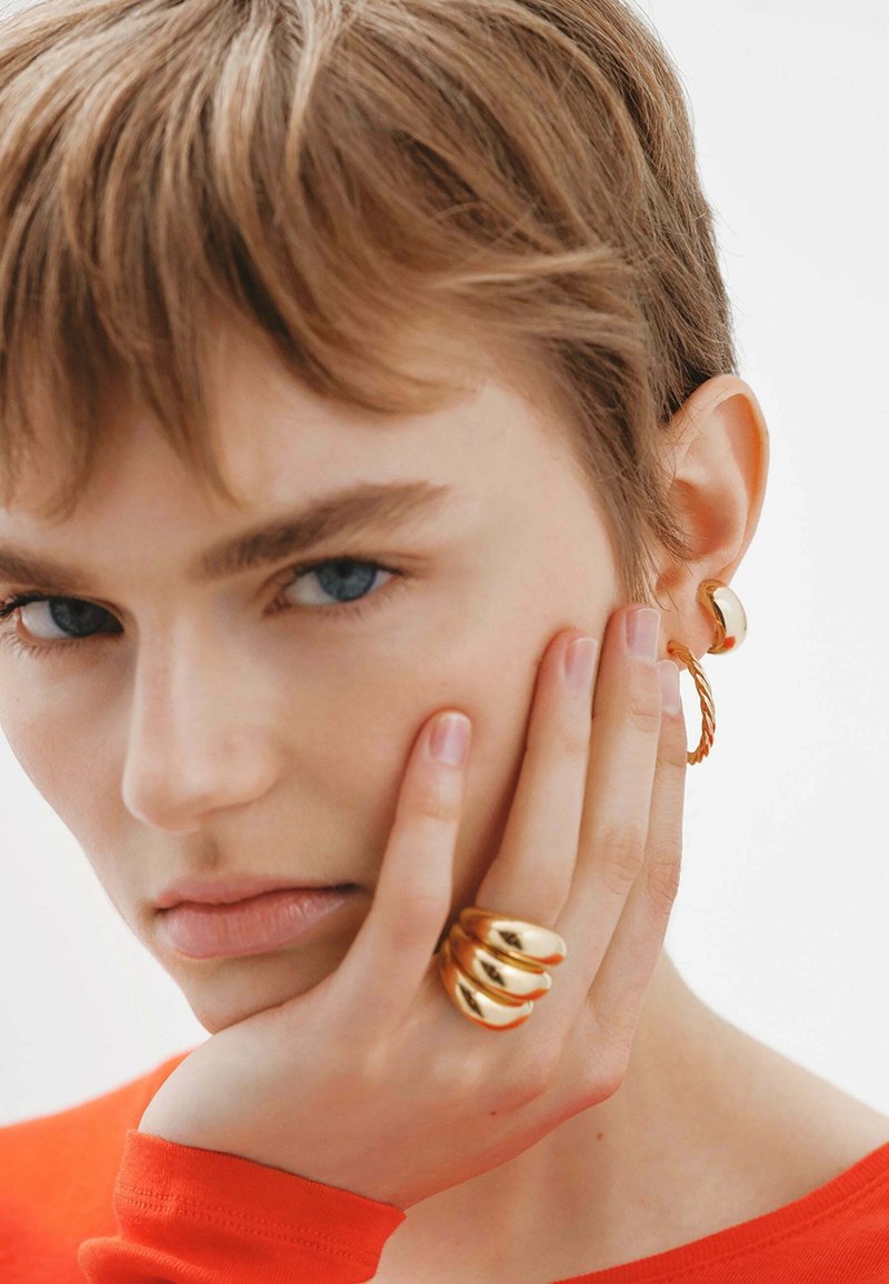 Pendientes de oro y un anillo de oro de tres aros, con superficies lisas y reflectantes, combinados con una blusa roja de manga larga. Vista en primer plano.