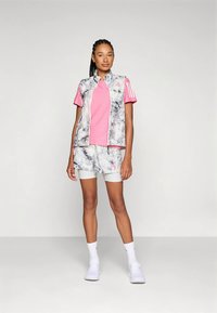 Sieviete stāv ar rozā krekliņu, baltu marmora raksta vestīti un šortiem, baltām kompresijas biksēm, baltām zeķēm un baltām sporta apaviem.