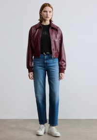 Μπορντό δερμάτινο μπουφάν τύπου bomber πάνω από μαύρο πουκάμισο, συνδυασμένο με ανοιχτό γαλάζιο τζιν κανονικής γραμμής και λευκά αθλητικά παπούτσια. Απλός σχεδιασμός, με ελάχιστες λεπτομέρειες.