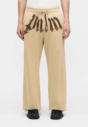 Osoba nosząca beżowe spodnie z szerokimi nogawkami z czarnym gotyckim napisem na górnej przedniej części oraz czarne trampki, stojąca na białym tle.