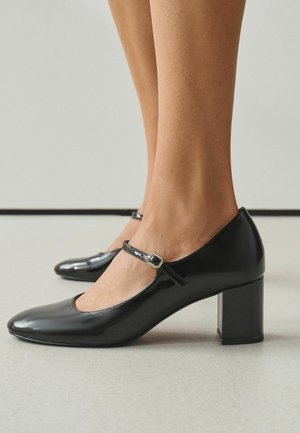 Beine, die glänzende schwarze Mary Jane Schuhe mit Blockabsätzen und Knöchelriemen tragen, stehen auf einem hellgrauen Boden.