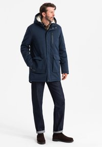 Parka blu navy con cappuccio, dotata di foderatura in pile, grandi tasche anteriori e tasca sul petto con zip. Abbinata a jeans scuri e stivali marroni.