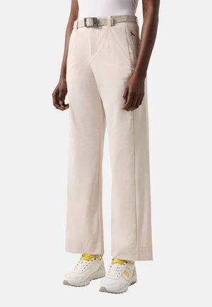 Osoba nosząca beżowe spodnie o szerokich nogawkach z boczną kieszenią na zamek, pleciony beżowy pasek oraz białe tenisówki z żółtymi sznurowadłami i detalami.