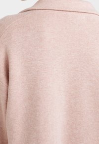 Světle růžový pletený svetr s texturovaným povrchem a klasikou límcem, zobrazující hladký, jemný povrch a decentní barevnou variaci.