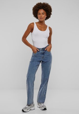 Hvit tanktopp, høyt liv blå jeans, avslappet passform, lyse vask, rette ben, brukt med sølvgrå sneakers. Enfarget bakgrunn.