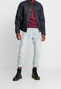 Giacca di jeans scura, felpa bordeaux, jeans azzurri con risvolti e stivali neri con lacci. Caratteristiche includono bottoni e tasche frontali.