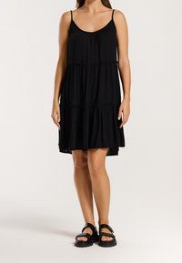 Femme portant une robe noire sans manches à volants et des sandales noires épaisses, debout devant un fond clair uni.