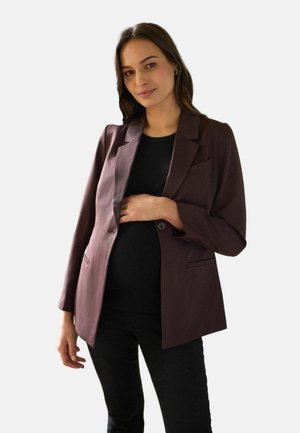 Kvinde iført en mørkebrun blazer over en sort top og bukser, der blidt holder sin gravide mave med den ene hånd.