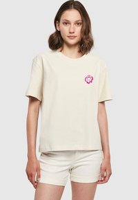 Krem bomull t-skjorte med rund hals og korte ermer som har en rosa tegneserie-lignende logo på venstre brystområde.