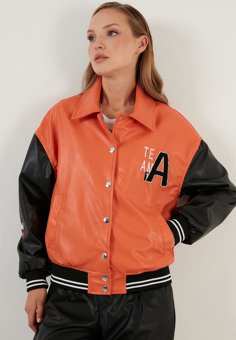 Oranje nep leren bomberjack met zwarte mouwen, voorzien van een knoopsluiting, gestreepte boorden en een geborduurd "TEAM A" logo.