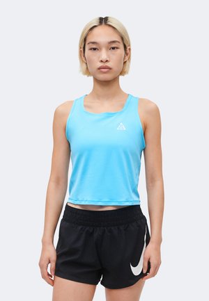 Persoană tânără cu păr scurt blond, purtând un top bleu deschis fără mâneci și pantaloni scurți negri Nike, pe fundal simplu.