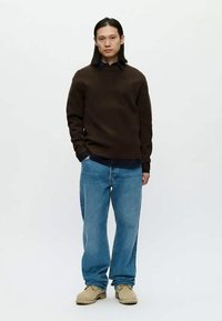 Mann som står med hånden i lommen, iført mørkebrun genser, mørk skjorte, løse blå jeans og beige sko mot hvit bakgrunn.