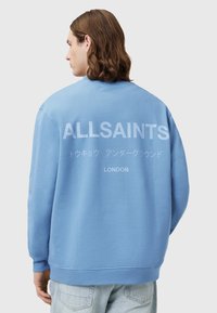 Světle modrý svetr s kulatým výstřihem, na zadní straně s velkým bílým nápisem "ALLSAINTS LONDON" a měkkou, texturovanou tkaninou.