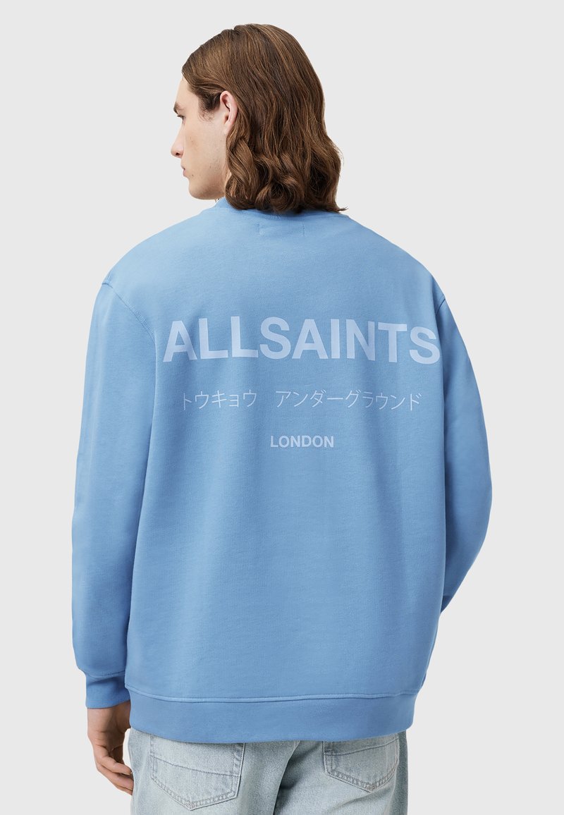 Světle modrý svetr s kulatým výstřihem, na zadní straně s velkým bílým nápisem "ALLSAINTS LONDON" a měkkou, texturovanou tkaninou.