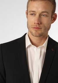 Zwarte blazer met scherpe revers over een wit overhemd. Voorzien van een klein accent van de Britse vlag op de linkerkraag. Gladde stofstructuur.