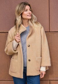 Beige wollen-blend jas met een gestructureerd visgraatpatroon, grote zakken en een enkele kraag, gecombineerd met een grijze trui en denim.